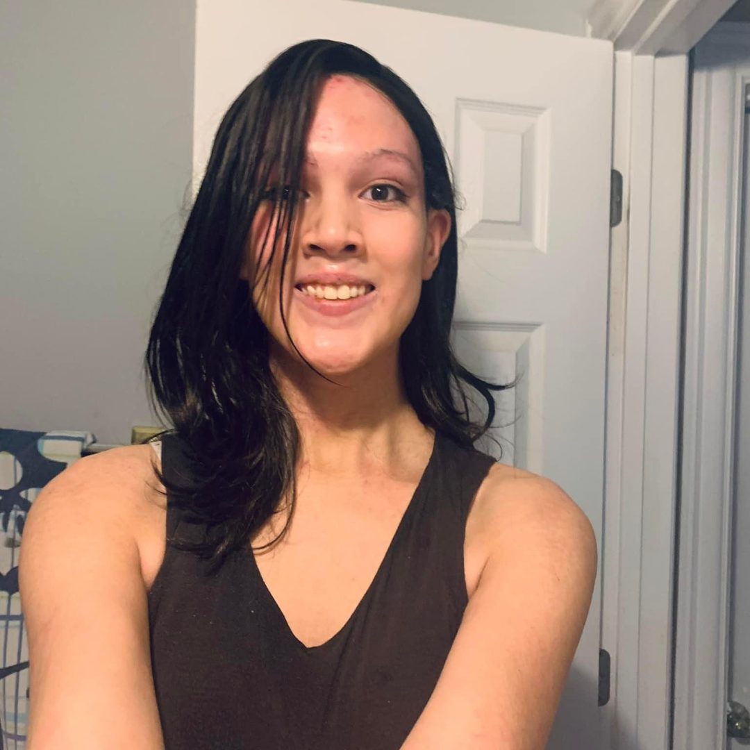 Photo of a smiling young trans woman of Asian-American descent with long dark hair standing in front of a white door. She is wearing a black tank top. 