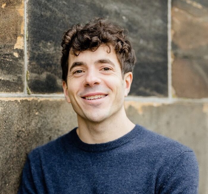 Photo of a white man with short curly brown hair and blue crew neck shirt.