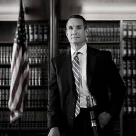whiteman in a dark suit leaning against a desk. there is a set of flags in the backgroundn. The man has a lightsaber on his hip.