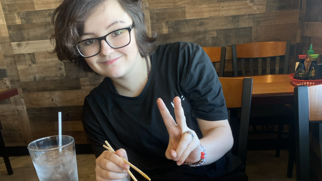 White teenager sitting in a restaurant holding chopsticks and showing the peace sign. Teen wears dark tee shirt and dark rimmed glasses. Teen has medium length wavy brown hair. Nonbinary.