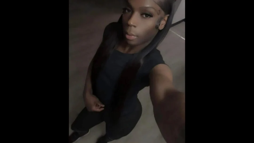 self portrait of a Black trans woman wearing a black short sleeve top and black leggings. Her hairs is shoulder length. she is looking at the camera with a serious expression. 